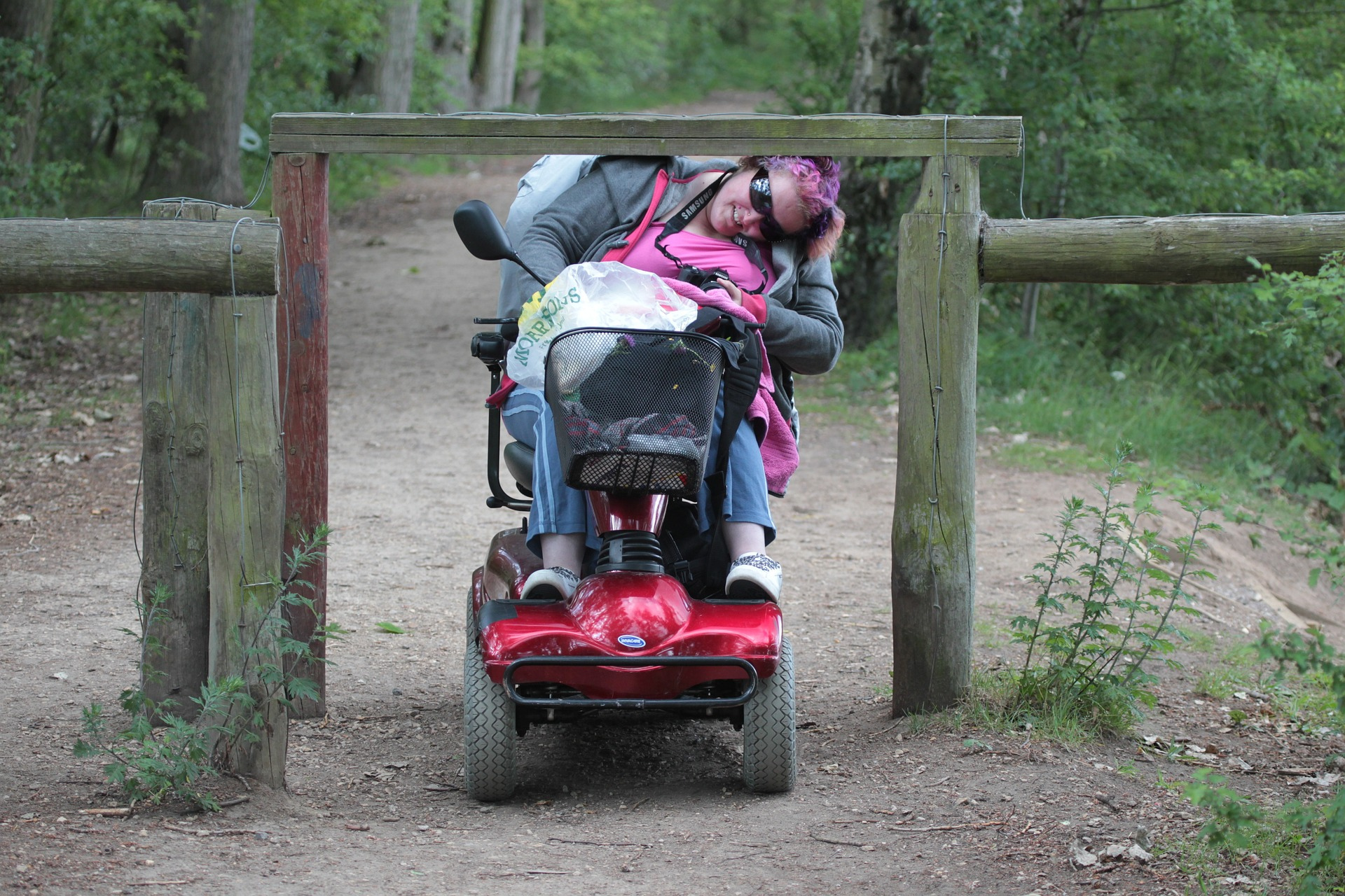 Vrouw in scootmobiel rijdt in bos. Zij kan niet onder de slagboom door.